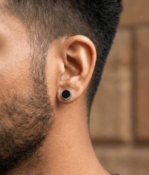 Close-up of a man's ear wearing a circular black stud earring with a silver rim, featuring short dark facial hair and a blurred brick background.