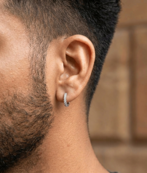 Side view of a man with a dark beard wearing a small silver hoop earring encrusted with clear diamonds or crystals against a blurred brick background.