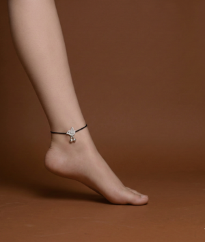 A high-quality photo of a fair-skinned person's foot and lower leg, standing on tip-toe against a solid brown studio background. The foot wears a simple black cord anklet featuring a detailed silver bird-shaped charm, specifically a dove.